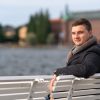 Attractive young man in warm coat and scarf sitting on a bench outdoors overlooking choppy water turning to smile at the camera