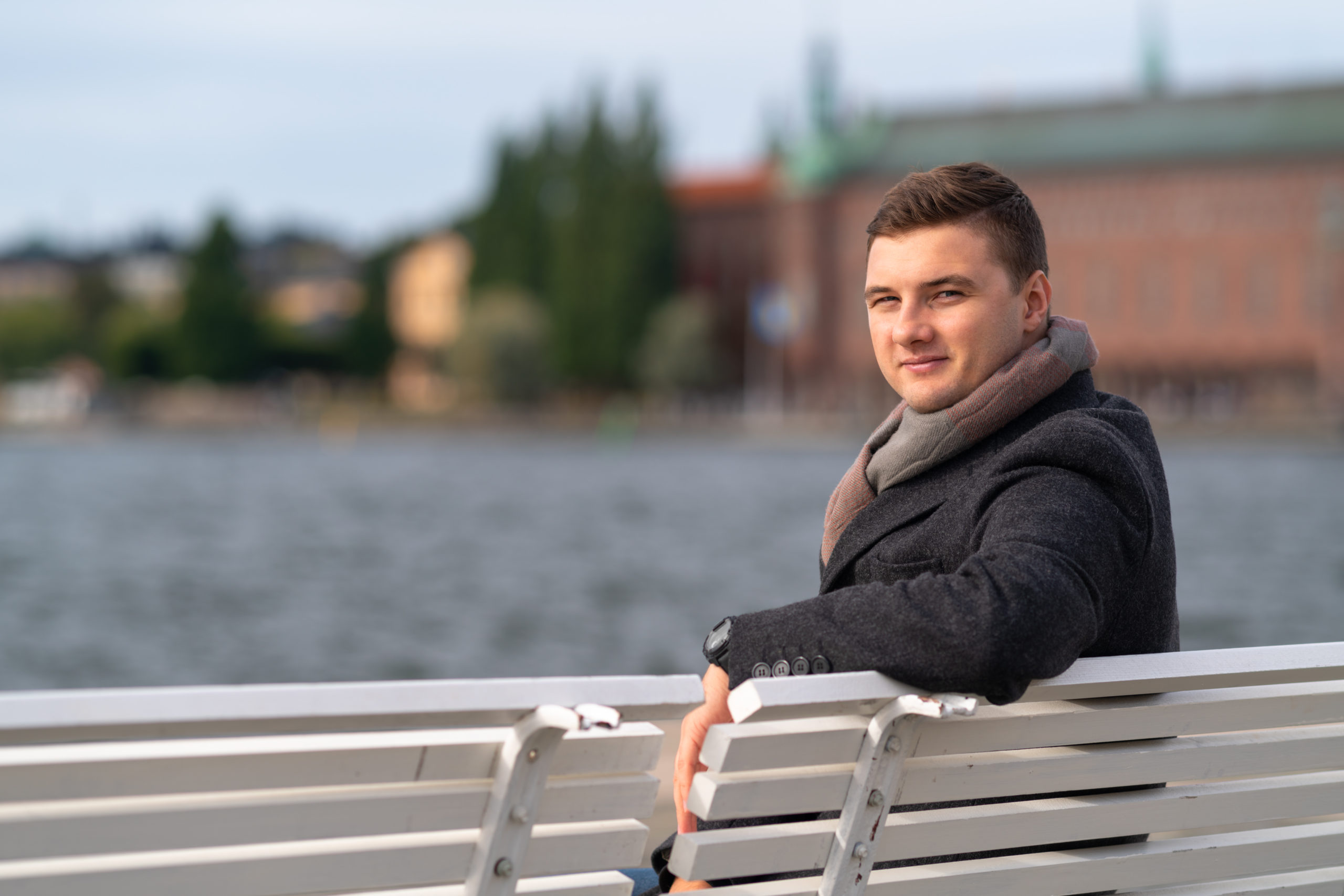Attractive young man in warm coat and scarf sitting on a bench outdoors overlooking choppy water turning to smile at the camera